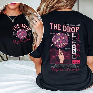 May include: Black t-shirt with a graphic design featuring a disco ball and text that reads "THE DROP" and "CRESCENT CITY". The design also includes the words "The White Raven" in a script font.