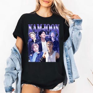 May include: Black t-shirt featuring a graphic of a group of people with the text "NAMJOON" in a purple, stylized font. The graphic has a vintage, faded look. The t-shirt is a classic crew neck style.