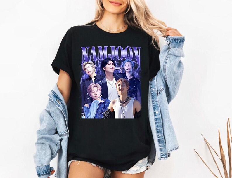 May include: Black t-shirt featuring a graphic of a group of people with the text "NAMJOON" in a purple, stylized font. The graphic has a vintage, faded look. The t-shirt is a classic crew neck style.