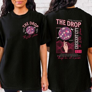 May include: Black t-shirt with a front and back graphic design. The front features a disco ball graphic with the text "THE DROP" and "The White Raven". The back has a disco ball graphic, text "THE DROP", "CRESCENT CITY", and "through love, all is possible."