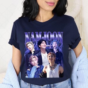 May include: Navy blue t-shirt featuring a graphic of five people with the name "NAMJOON" in a purple, retro font. The graphic has a vintage aesthetic with lightning bolt accents. The shirt is a crew neck.