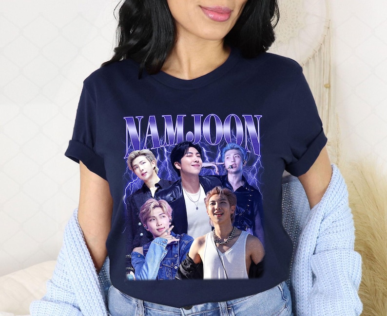 May include: Navy blue t-shirt featuring a graphic of five people with the name "NAMJOON" in a purple, retro font. The graphic has a vintage aesthetic with lightning bolt accents. The shirt is a crew neck.