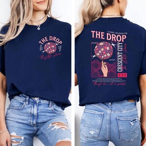 May include: Navy blue t-shirt with a graphic featuring a disco ball design and the text "THE DROP". The back of the shirt includes the text "COME CELEBRATE THE DROP CRESCENT CITY" and a hand balancing a disco ball.