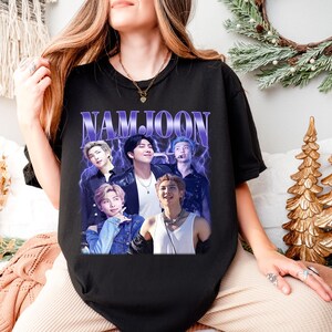 May include: Black t-shirt featuring a graphic of a group of people with the text "NAMJOON" in a purple, retro font. The graphic has a vintage, faded look. The shirt is a crew neck style.