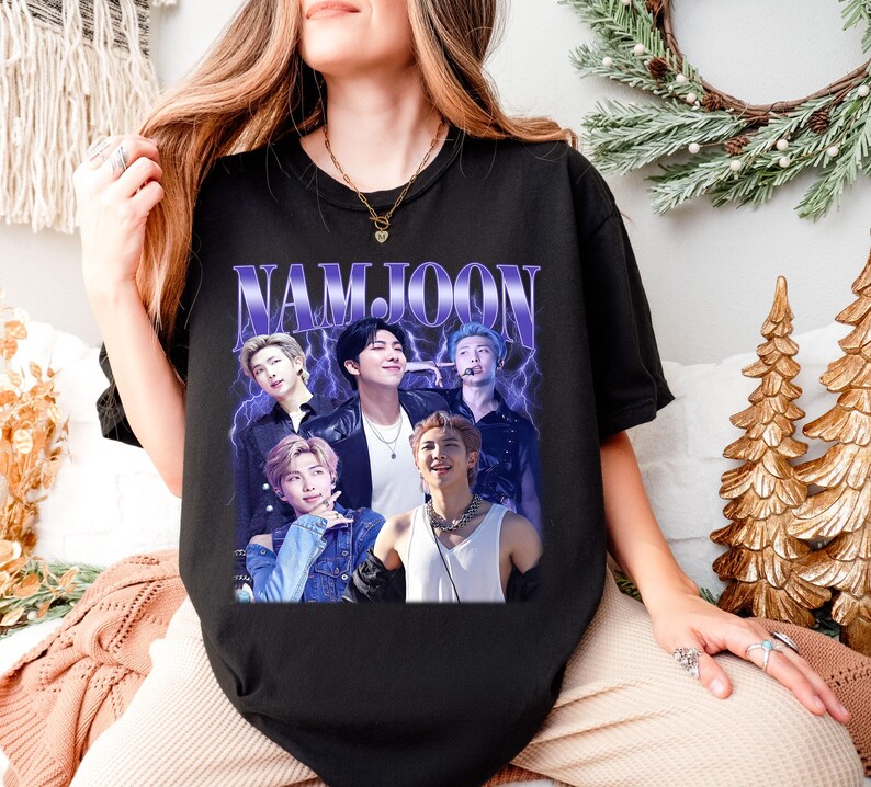 May include: Black t-shirt featuring a graphic of a group of people with the text "NAMJOON" in a purple, retro font. The graphic has a vintage, faded look. The shirt is a crew neck style.