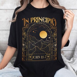 May include: Black t-shirt with a gold graphic design. The design features the words "IN PRINCIPIO" and text from John 1:1-7, along with a mountain and moon illustration. The shirt is a classic crew neck.