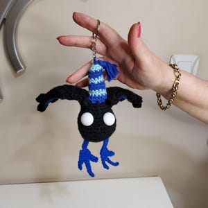 May include: A handmade crochet keychain featuring a black creature with two large white circular eyes, blue feet, and a blue and light blue striped pointed hat topped with a small blue tassel. The figure is attached to a silver metal keyring.
