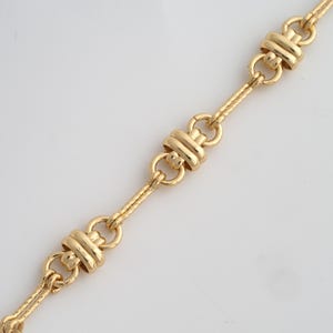 May include: Gold-toned necklace with a repeating pattern of linked segments. Each segment features a central, cylindrical design flanked by circular links. The chain has a textured, woven appearance, creating a delicate yet structured aesthetic.
