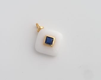 May include: A white pendant with a gold bail and a square, dark blue gemstone set in a gold frame. The pendant has a rounded square shape and is likely made of a smooth, opaque material.