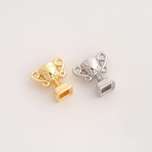 May include: Two small trophy charms, one finished in a polished gold tone and the other in a polished silver tone. Each trophy features two curved handles, a textured bowl section with a floral pattern, and a square base with a hollow centre.