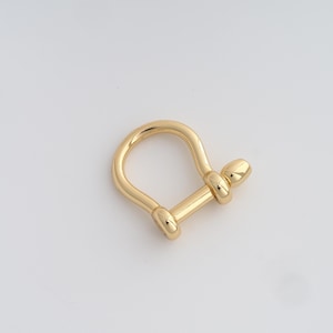 Peut inclure: Un fermoir en forme de manille en or. Le fermoir a un dessus arrondi et une barre droite avec des extrémités arrondies. Le métal a une surface brillante et réfléchissante. Le fermoir est conçu pour la fabrication de bijoux ou d'autres projets d'artisanat.