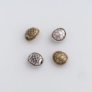 May include: Four metal beads in a variety of finishes. Two are gold-toned, one with a geometric pattern and the other with a honeycomb design. The other two are silver-toned, one with a geometric pattern and the other with a honeycomb design. Each bead has a small hole.
