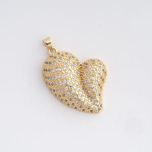May include: A gold-coloured heart-shaped pendant with a bail at the top. The pendant is covered in small, sparkling clear and gold-coloured stones. The heart shape is slightly asymmetrical, with a rounded top and a more pointed bottom.
