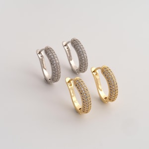May include: Two pairs of hoop earrings, one pair in silver-tone metal and one pair in gold-tone metal. Each hoop features a curved, thick profile encrusted with numerous small, clear, sparkling stones along the entire outer surface of the hoop.