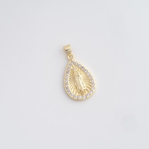 May include: Gold-colored pendant in a teardrop shape, featuring a religious figure in the center. The pendant is surrounded by a halo of small, clear crystals. The pendant is on a white background.