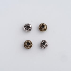 May include: Four decorative metal beads. Two are silver-toned, and two are bronze-toned. Each bead has a floral design with small circles around the center hole. The beads are round and have a hole through the center for stringing.