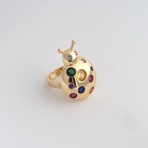 Könnte beinhalten: Ein goldfarbener Marienkäfer-Ring mit bunten Edelsteinen. Der Ring hat einen abgerundeten Körper mit verschiedenfarbigen Edelsteinen, darunter grün, gelb, blau und rot. Der Kopf des Marienkäfers hat einen klaren Edelstein und zwei Fühler.