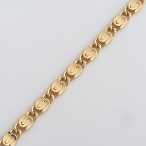 Può includere: Una collana a catena color oro con un design a maglie ovali ripetute. I collegamenti sono collegati da connettori curvi più piccoli, creando un motivo continuo. La catena è esposta su uno sfondo bianco.