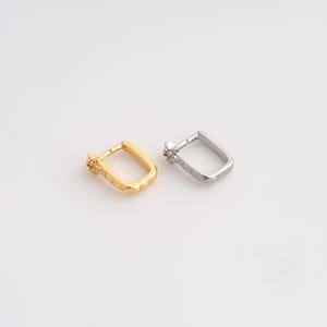 May include: Two small rectangular hoop earrings, one in a polished gold tone and one in a polished silver tone. Each earring features a textured, geometric pattern along the outer edge and a small, decorative cluster of tiny stones at the hinge.