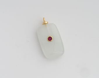 Puede incluir: Un colgante rectangular con la parte superior e inferior redondeadas, con una gema roja central engastada en un bisel dorado. El colgante tiene una argolla dorada y está hecho de un material translúcido blanquecino.