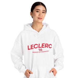Może przedstawiać: Biała bluza z kapturem z napisem "LECLERC 16 PRINCE OF MONACO" w kolorze czerwonym. Bluza ma kaptur ze sznurkiem i przednią kieszeń. Modelka ma na sobie bluzę.