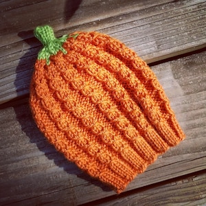 Könnte beinhalten: Orangefarbener gestrickter Kürbis-Hut mit grünem Stiel. Der Hut ist mit einem strukturierten Stichmuster gearbeitet.