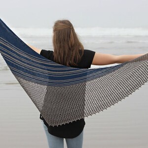 Könnte beinhalten: Eine Frau trägt einen blau-grauen gestreiften Schal mit einem Spitzenmuster. Der Schal ist über ihre Schultern gelegt und sie steht am Strand.