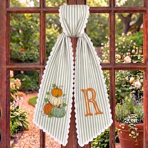 May include: A hanging fabric door swag featuring a light gray and white vertical striped pattern with white scalloped lace trim. One side displays an embroidered cluster of three pumpkins in orange, white, and green, while the other side features a large orange embroidered letter R.