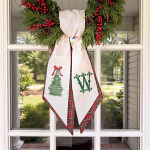 Peut inclure: Une couronne de fête composée de branches de cèdre vertes, de pommes de pin et de baies rouges, avec un ruban en tissu crème suspendu. Le ruban présente un sapin de Noël brodé en vert avec un nœud rouge et une grande lettre W brodée en vert.