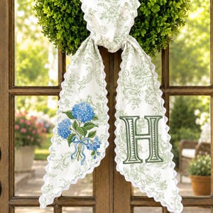 May include: A white fabric door scarf with a green floral pattern and scalloped edges. It features a blue hydrangea bouquet with a blue ribbon and the letter "H" embroidered in green. The scarf is hanging on a wooden door.