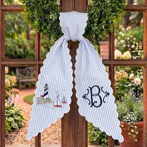 May include: A light blue and white striped fabric door hanger with scalloped edges. The left side features an embroidered lighthouse and two sailboats. The right side displays a large, dark blue embroidered letter B with decorative flourishes.