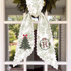 Peut inclure: Une longue écharpe en tissu blanc avec un motif toile de Jouy vert clair et des bords festonnés. Le côté gauche présente un sapin de Noël brodé en vert avec un nœud rouge, et le côté droit affiche une lettre H rouge dans une couronne verte.