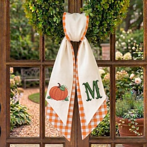 May include: A decorative fabric door hanger featuring a cream-colored base with orange and white gingham trim. One side displays an embroidered orange pumpkin with green leaves, while the other side features a large, embroidered green letter M.