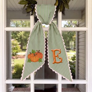 May include: A green and white striped fabric door hanger with scalloped edges. The left side features an embroidered graphic of three orange pumpkins with green leaves, and the right side features a large, embroidered orange letter E.