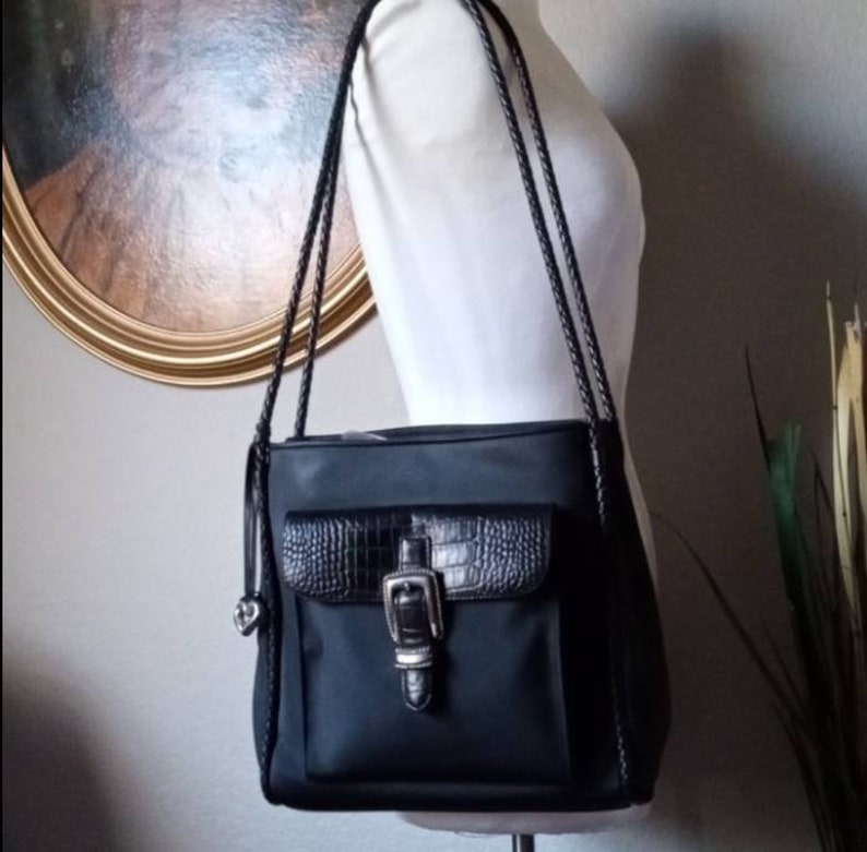 Może przedstawiać: Czarna torebka na ramię z kieszenią z krokodylej sk&oacute;ry i srebrną klamrą. Torebka ma pleciony pasek.