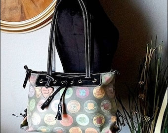 May include: A multi-colored tote bag with a black leather trim and handles. The bag has a drawstring closure and a heart-shaped charm with the text "Tommy &  Heidi" on it. The bag is covered in colorful circles with different designs and text.