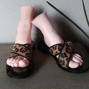 May include: A pair of black leather sandals with a leopard print strap and gold buckle. The sandals have a criss-cross strap design.