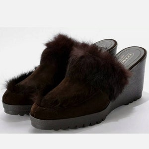 May include: Brown suede wedge heeled mules with a fur trim around the top of the shoe. The shoes have a black rubber sole and the brand name "COACH" is visible on the side of the shoe.