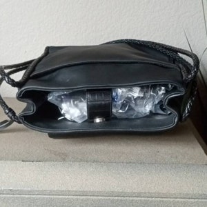 Może przedstawiać: Czarna sk&oacute;rzana torebka z plecionym paskiem i srebrną klamrą. Torebka jest otwarta i pusta.