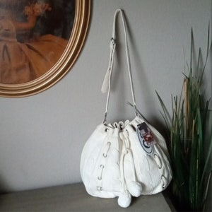 May include: White knitted drawstring bucket bag with a long strap and white pom poms. The bag has a cable knit pattern and silver eyelets.