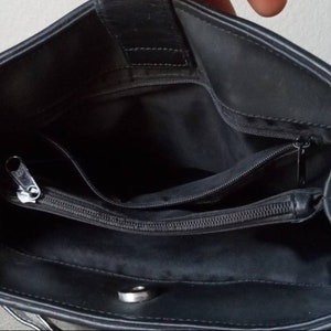 Może przedstawiać: Czarna sk&oacute;rzana torebka z kieszenią zapinaną na zamek błyskawiczny i zatrzaskiem. Torebka jest otwarta i widać wnętrze.