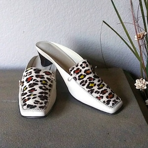 May include: A pair of white heeled mules with a leopard print design on the toe. The heels are about 3 inches tall.