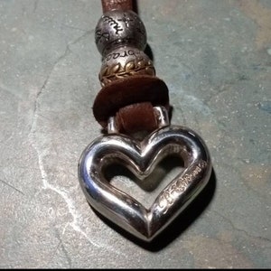 May include: A silver heart-shaped charm with a cutout heart and the word "Dreams" inscribed on it. The charm is attached to a brown leather cord with silver beads.