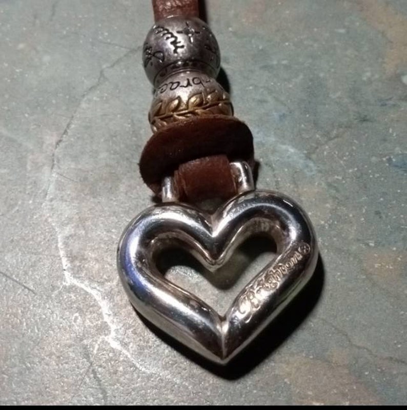 May include: A silver heart-shaped charm with a cutout heart and the word "Dreams" inscribed on it. The charm is attached to a brown leather cord with silver beads.