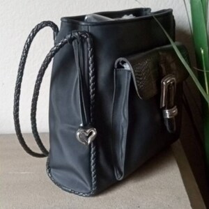 Może przedstawiać: Czarny sk&oacute;rzany plecak z plecionym paskiem i srebrnym wisiorkiem w kształcie serca. Plecak ma dużą kieszeń z przodu z srebrną klamrą.