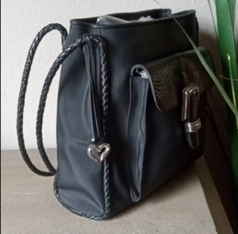 Może przedstawiać: Czarny sk&oacute;rzany plecak z plecionym paskiem i srebrnym wisiorkiem w kształcie serca. Plecak ma dużą kieszeń z przodu z srebrną klamrą.