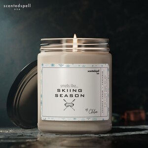 Puede incluir: Una vela encendida en un tarro de cristal con tapa negra. La etiqueta dice "smells like... SKIING SEASON" con un gráfico de esquí. El aroma es "Coconut Cream + Cardamom". La marca es "scentedspell USA".