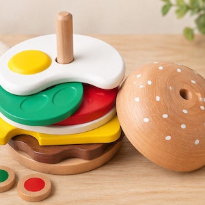 Puede incluir: Una hamburguesa de juguete de madera con varios componentes coloridos. La hamburguesa incluye una parte superior blanca con puntos blancos, una clavija de madera y capas de ingredientes coloridos como un huevo frito, lechuga, tomate y una hamburguesa. También hay dos pequeños círculos de madera con puntos verdes y rojos.