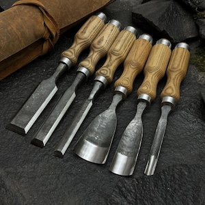 Puede incluir: Un juego de seis cinceles de madera con mangos de madera y hojas de metal, dispuestos sobre una superficie oscura. Los cinceles varían en forma y tamaño de hoja, con un rollo de herramientas de cuero en el fondo. Los mangos tienen un patrón de veta de madera clara.