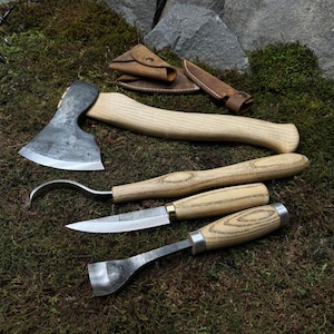 Puede incluir: Un conjunto de herramientas para trabajar la madera, que incluye un hacha pequeña, un cuchillo curvo, un cuchillo recto y una gubia, con mangos de madera y hojas de metal. También hay una funda de cuero y una pequeña bolsa de cuero. Las herramientas están dispuestas sobre musgo verde.